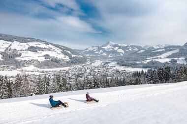 Zwei Menschen rodeln auf einer verschneiten Piste mit Blick auf die winterliche Berglandschaft.