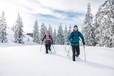 Zwei Personen beim Schneeschuhwandern durch verschneite Winterlandschaft im Skigebiet, verschneite Tannen rechts.
