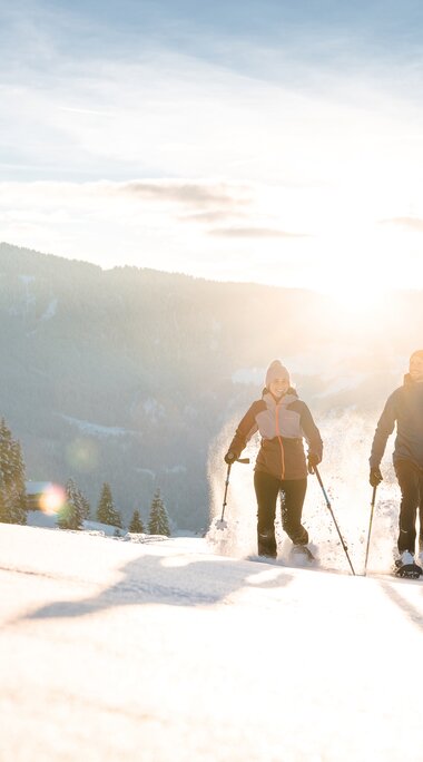 Zwei Personen gehen mit Schneeschuhen über eine verschneite Wiese, Sonnenaufgang und Berge im Hintergrund.