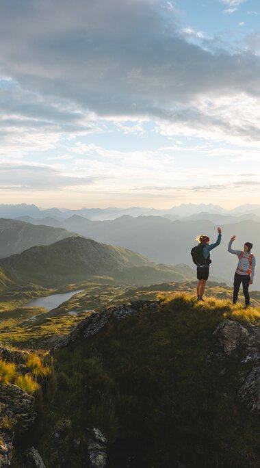 Zwei Wanderer stehen auf einem Bergkamm bei Sonnenaufgang in den Alpen.