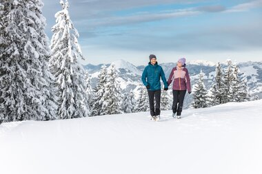 Zwei Winterwanderer laufen im Schnee durch verschneite Tannen vor den Bergen im Hintergrund.
