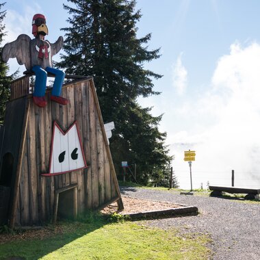 Holzfigur mit markiertem Gesicht und Flügeln auf einem Spielplatz im Alpengebiet, im Hintergrund Berge und Bäume.