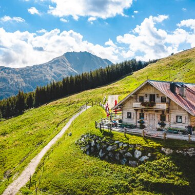 Almhütte auf einer grünen Wiese mit Bergpanorama in den Alpen bei sonnigem Wetter.