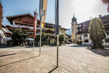Der Dorfplatz von Westendorf mit Pfarrkirche, traditionellen Gebäuden und Aufenthaltsbereichen im Ortszentrum.