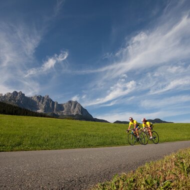 Zwei Rennradfahrer fahren auf einer Straße vor dem Panorama des Wilden Kaisers in der Region St. Johann in Tirol