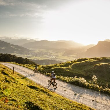 Zwei Mountainbiker auf einer Höhenroute oberhalb der Region St. Johann in Tirol mit weitem Blick über das Tal und die umliegende Bergwelt