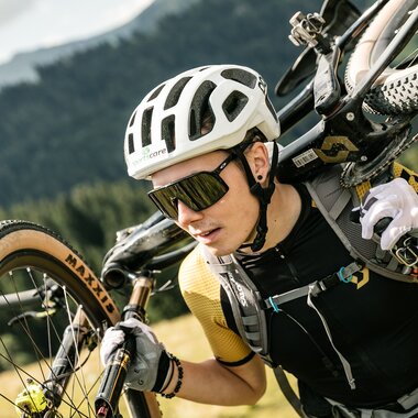 Mountainbiker trägt sein Fahrrad über die Schulter auf einem alpinen Weg in der Region St. Johann in Tirol und trägt dabei Helm, Rucksack und Sportbekleidung