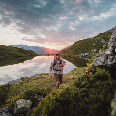 Wanderer mit Rucksack an einem ruhigen Bergsee in der Kelchsau bei Sonnenuntergang, umgeben von grünen Berghängen und Felsen.